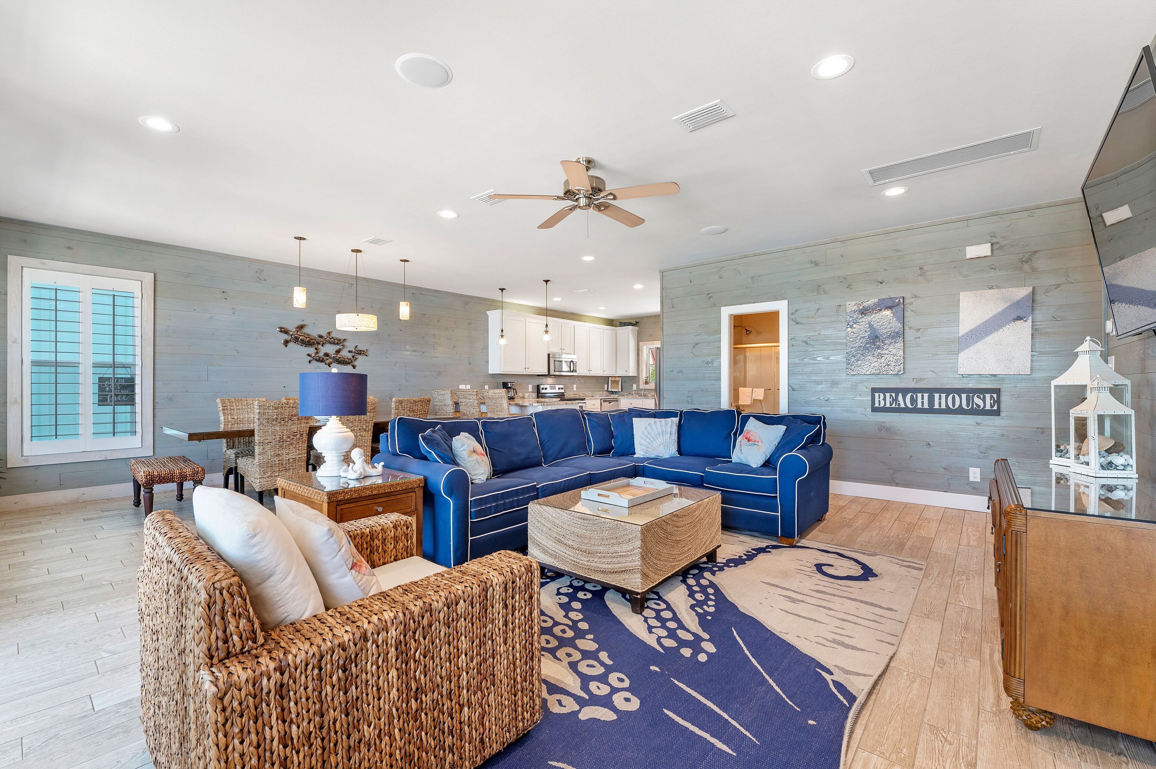 Un salón amplio con un sofá seccional azul, muebles de mimbre, un ventilador de techo y un cartel en la pared que dice 'Beach House'.