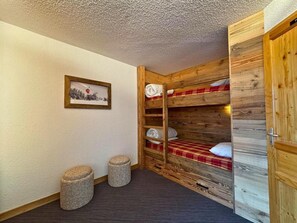 A room with a bunk bed, a small stool, a framed picture on the wall, and a wooden door.