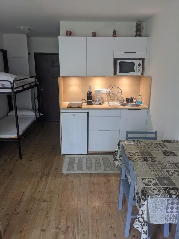 A compact kitchen with white cabinets, a microwave, and a coffee maker. There is a bunk bed to the left and a dining table with a patterned tablecloth to the right.