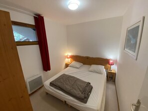A single bed with a headboard, two bedside lamps, a window with red curtains, a radiator, and a framed picture on the wall. - 200m from ski slopes, 2nd floor, view mountain, balcony, tv, 31m², Superdévoluy (Le Dévoluy)