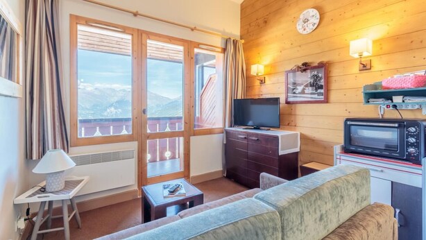 Ein gemütliches Zimmer mit Holzbalkendecke, einer Couch, einem Fernseher, einem kleinen Tisch und einem Fenster mit Vorhängen.