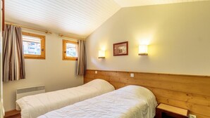 Ein Zimmer mit zwei Einzelbetten, einem Holzkopfteil, einem kleinen Nachttisch, einem Fenster mit Vorhängen und zwei Wandleuchten.