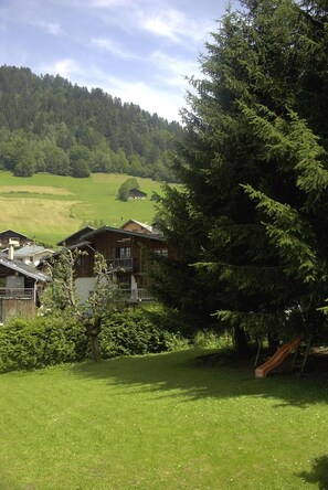 A wooden chalet with a balcony, surrounded by lush greenery and a forested hillside.