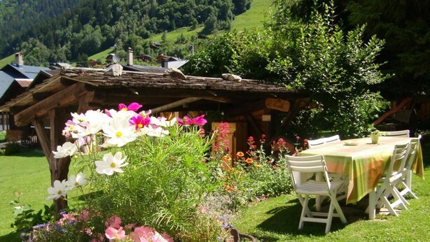 Eine rustikale Holzstruktur mit einem Garten davor, einem Tisch mit Stühlen und einer Bergkulisse im Hintergrund.