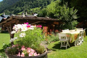 A rustic wooden structure with a garden in front, a table with chairs, and a mountainous backdrop.