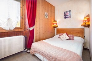 A bedroom with a wooden headboard, a window with a white curtain, a radiator, and two wall-mounted lamps.