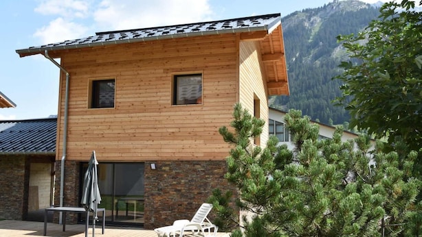 Une maison en bois à deux étages, dotée d’une façade en pierre, d’un patio avec une table et des chaises, et offrant une vue sur les montagnes en toile de fond.