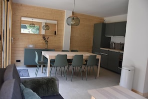 A modern kitchen with dark cabinets, a dining area with a white table and green chairs, and a wooden wall with a window.