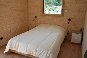 A bedroom with a wooden floor, a bed with white bedding, a nightstand with a drawer, and a window with a view of parked cars and greenery.