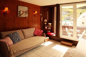 A wooden-furnished room with a sofa, a wall-mounted lamp, a poster, and a view of outdoor seating through a glass door.