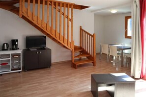 A staircase with wooden railings leading to an upper floor, a television on a cabinet, a dining area with chairs and a table, and a small table with a metal frame.