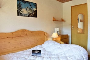 A hotel room with a wooden headboard, a bed with white bedding, a bedside table with a lamp, a coffee maker, a framed picture on the wall, and a wooden door with a mirror. - 50m from shops, 1st floor, balcony, tv, 18m², Méribel (Méribel - Les Allues)