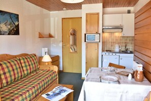 A cozy room with a checkered sofa, a small table with a newspaper, a wooden door, a kitchenette with a stove and microwave, and a painting on the wall. - 50m from shops, 1st floor, balcony, tv, 18m², Méribel (Méribel - Les Allues)