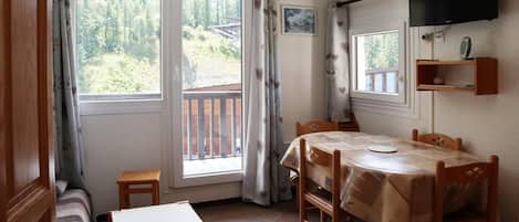 A room with a dining table, chairs, a small table, a sofa, a television mounted on the wall, and a window with curtains.
