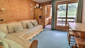 A cozy room with a wooden sofa, a dining table, and a view of a garden through a large window. - 10m from ski slopes, 4th floor, 27m², Méribel-Mottaret (Méribel - Les Allues)