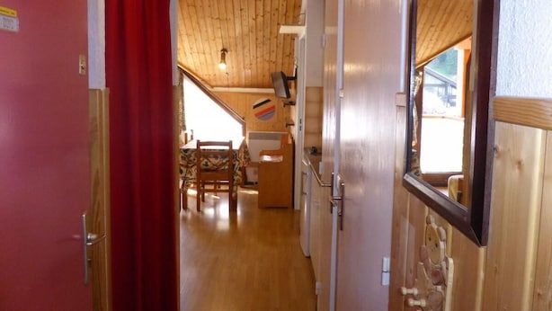 Un pasillo estrecho con paredes de madera y una cortina roja a la izquierda. Se ve en el fondo un techo de madera y una pequeña zona de cocina con una mesa y sillas.