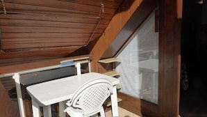 Un pequeño balcón cerrado con una mesa y una silla blancos, techo de madera y una ventana con cortina.