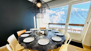 En matplats med ett runt bord för sex personer, vita stolar och utsikt över snöklädda berg genom stora fönster.