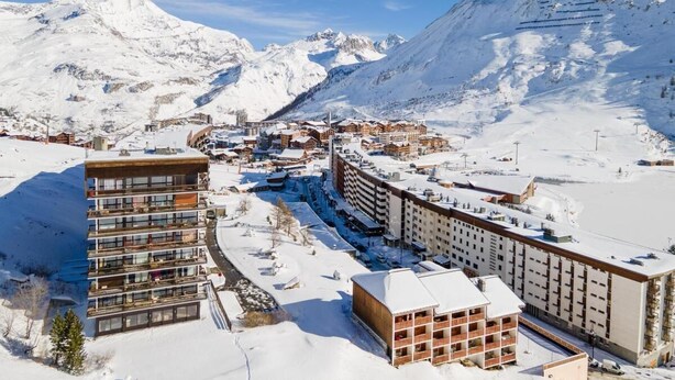 En snötäckt bergsby med moderna byggnader, inklusive ett hotell med flera våningar och balkonger, och en skidort i bakgrunden.