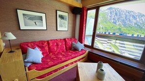 A room with a red sofa, a wooden table, and a window with a mountain view. - 10m from ski slopes, 4th floor, balcony, tv, ski locker, 52m², Tignes (Tignes)