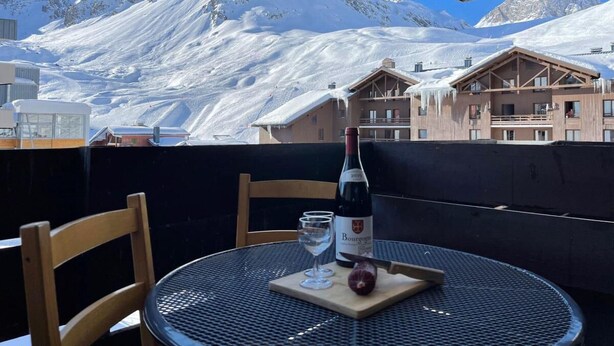 Ein Tisch mit einer Weinflasche, Gläsern und einem Holzbruch mit einem Apfel, vor einer verschneiten Berglandschaft.