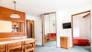 Et kompakt værelse med et træbord, stole, en rød sofa, et lille skab og et fjernsyn ophængt på væggen.