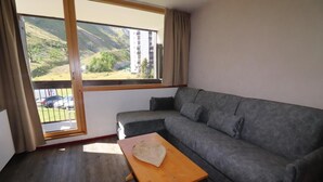 A modern living room with a grey sofa, a wooden table, and a large window offering a view of a building and greenery.