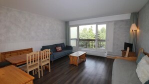A room with a wooden table, a sofa, a small wooden table, and a bed. There is a window with a view of trees and a balcony.