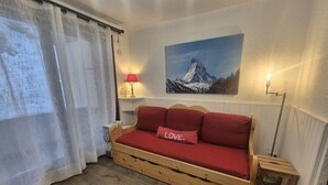 Une pièce chaleureuse avec un canapé-lit en bois, un coussin rouge orné du mot « AMOUR », un tableau représentant une montagne et une lampe sur une table de chevet.