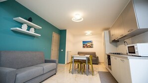 En kompakt opholdsstue med en grå sofa, et spisebord med stole, et køkkenområde med skabe og hårde hvidevarer, samt vægge i turkis.