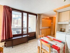 Uma cozinha pequena com armários de madeira, uma janela com cortinas, uma mesa de jantar com toalha e um radiador sob a janela.