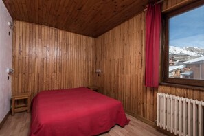Quarto com painéis de madeira, uma cama, uma janela com vista para edifícios cobertos de neve e um radiador.