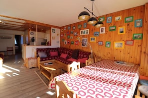 Una sala de estar acogedora con un sofá de madera, una mesa de comedor con mantel estampado y una zona de cocina al fondo.