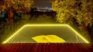 Terraza de madera con zona delimitada por una red y un cojín amarillo, rodeada de árboles y un paisaje urbano al fondo.