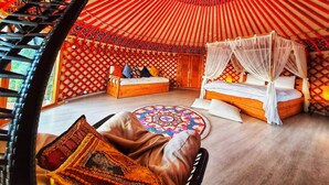 Una yurta tradicional con un tragaluz redondo, dos camas de madera con dosel, un tapete tejido y un sillón colgante.