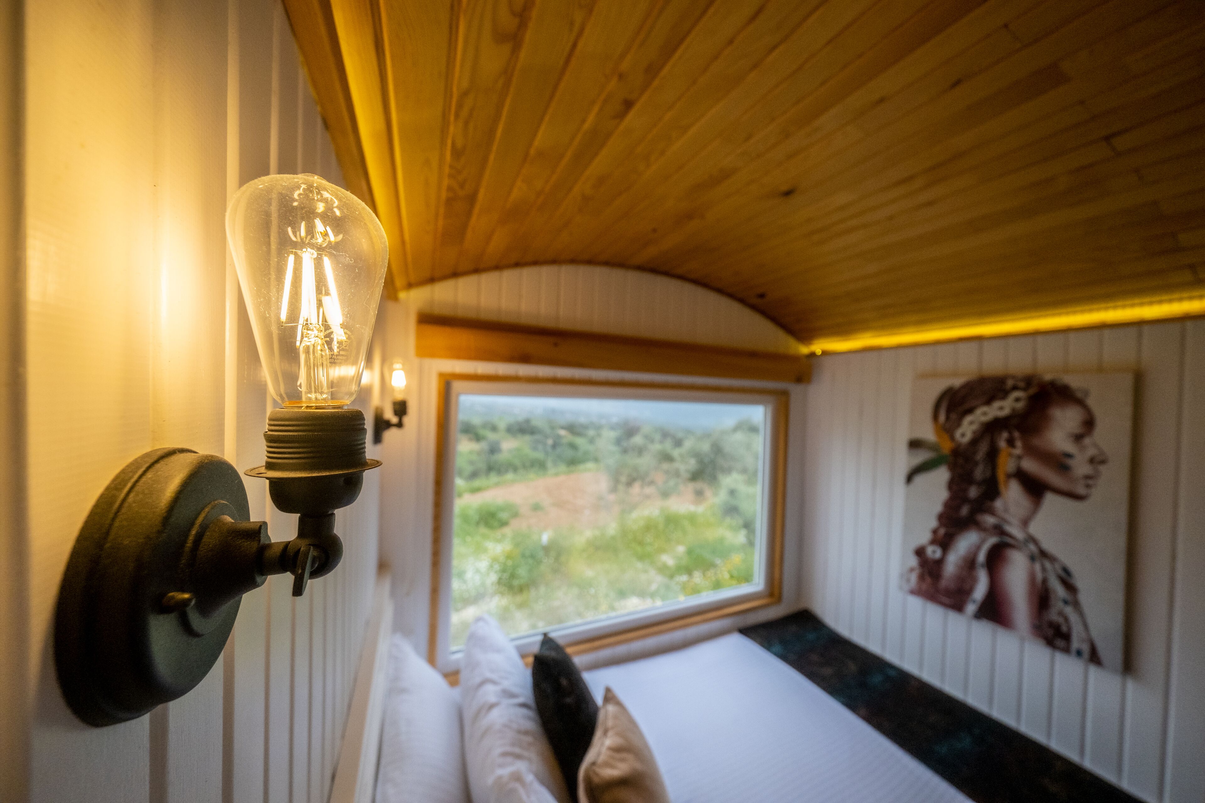 A room with a bed, a wall-mounted light fixture, a window with a view of greenery, and a painting on the wall.