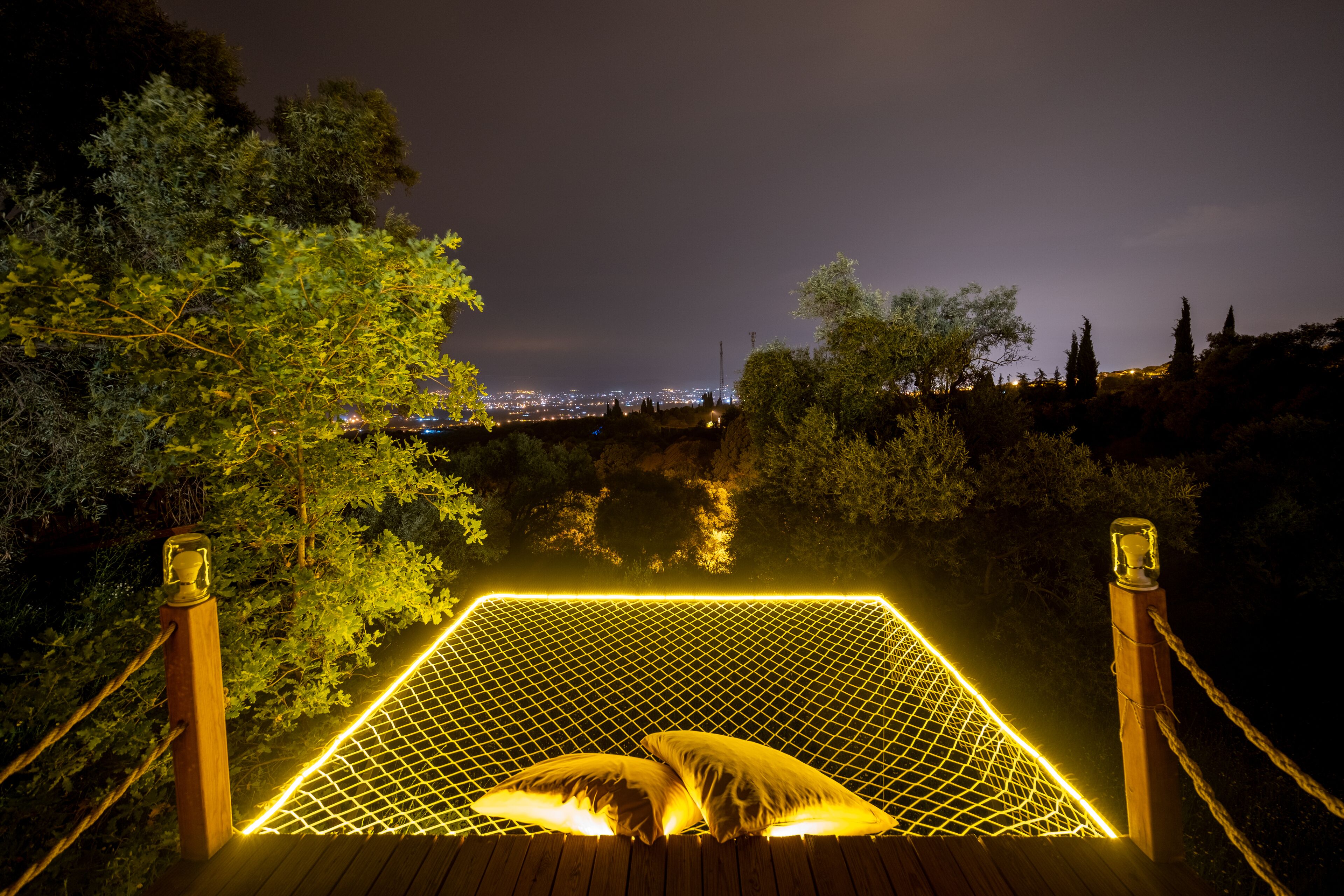 Una hamaca con superficie de mosquitero y dos almohadas, colocada en una terraza de madera con cuerdas a cada lado, con vista a un paisaje urbano nocturno.