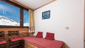 A room with a bed, two red pillows, a window with a view of snow-covered mountains, and a wooden chair with a red cushion. - 10m from resort center, 11th floor, television, ski locker, 12m², Tignes (Tignes)