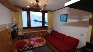 A cozy room with a wooden table, red sofa, and a view of a snowy landscape through large windows. - 10m from resort center, 11th floor, television, ski locker, 12m², Tignes (Tignes)
