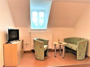 A small, cozy room with a sloped ceiling, a television on a wooden cabinet, two patterned armchairs, a small round table, and a window with a view. - Ferienzimmer Nr. 34 - Schullandheim Reichwalde (Boxberg/Oberlausitz)