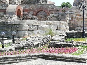 An ancient stone structure with a brick archway, stone steps, and a flower bed with pink flowers.