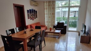 Een ruime woonkamer met een houten eettafel, zwarte stoelen, een bank en een salontafel. Er is een schuifdeur die leidt naar een balkon met uitzicht op groen.