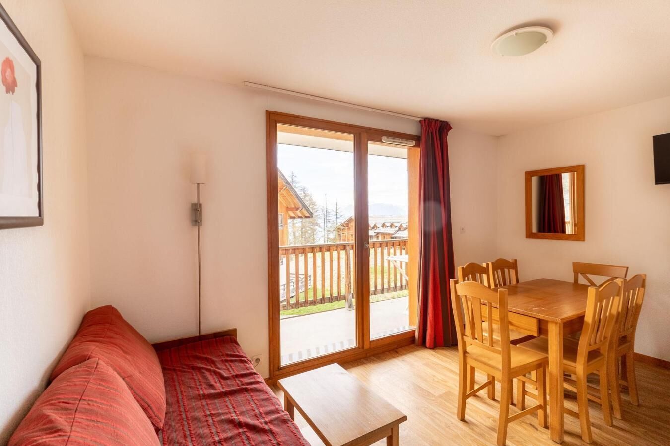 A room with a wooden table and chairs, a red sofa, and a sliding glass door leading to a balcony with a wooden fence.