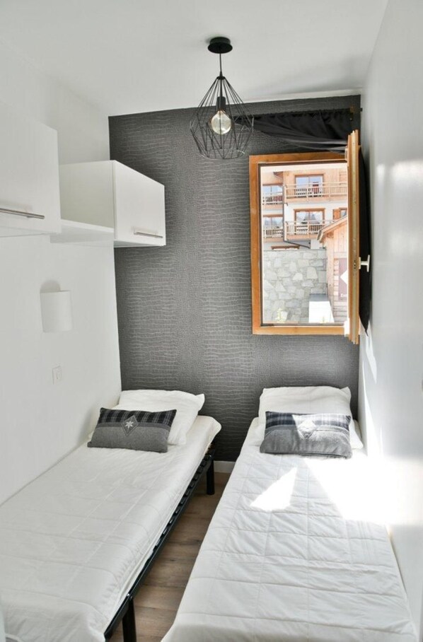 A small, modern room with two single beds, a large window with a view of a building, and a unique pendant light fixture.