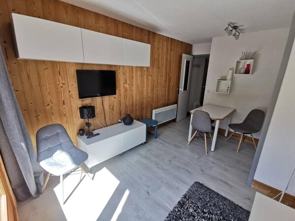 A modern living room with a wooden wall, a flat-screen TV, a sofa, a coffee table, and a dining area with chairs and a table.