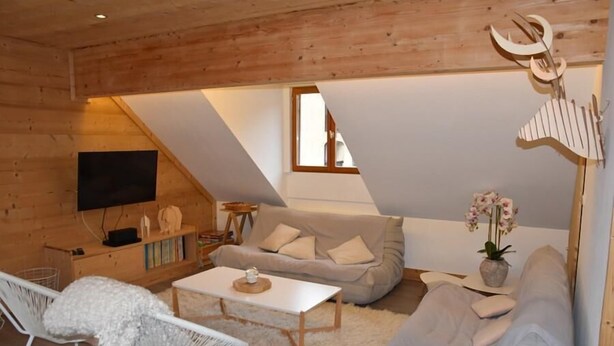 Ein gemütliches Wohnzimmer im Loft-Stil mit schrägem Dach, Holzwänden, einer Couch, einem weißen Sessel und einem flachbildfernseher an der Wand.