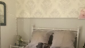 Ein Bett mit Metallgestell und gesteppter Bettdecke, ein Nachttisch mit Lampe sowie eine Wand mit ornamentalem Wandpapier.