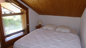 A bedroom with a wooden ceiling, a large window, a bed with white bedding, and a nightstand with a lamp.