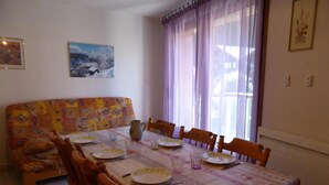 A dining area with a table set for four, surrounded by wooden chairs. A colorful sofa is against the wall, and there are two framed pictures on the wall.