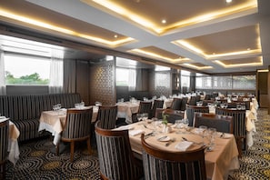 A spacious dining area with multiple tables and chairs, well-lit with ceiling lights, and large windows providing natural light.